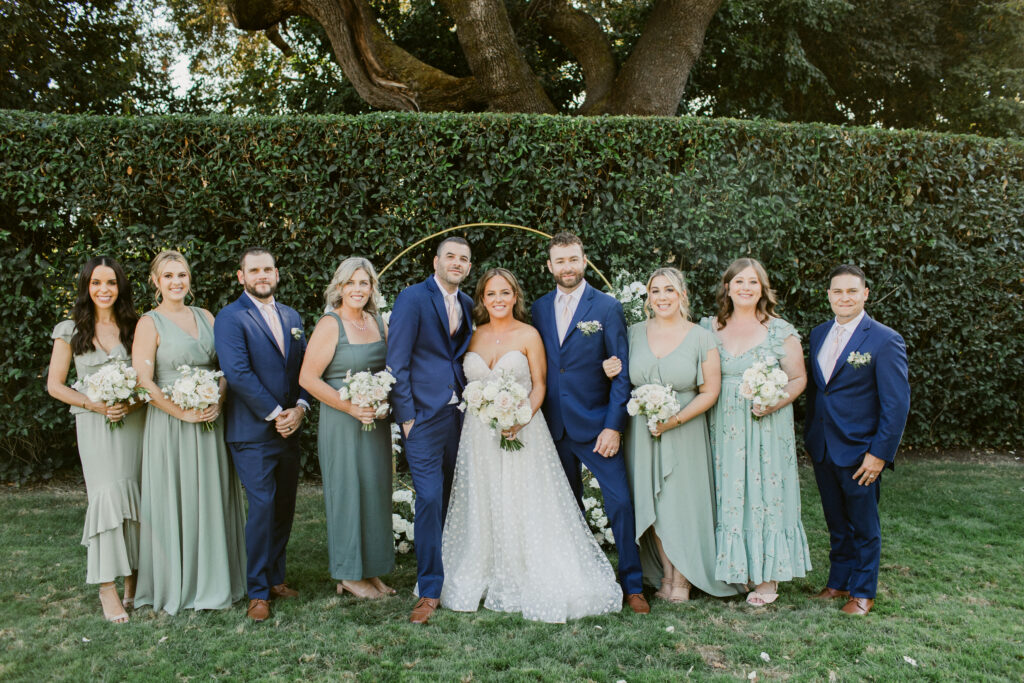 Wedding party dressed in coordinated sage green and blue attire, posing together in a scenic outdoor setting.